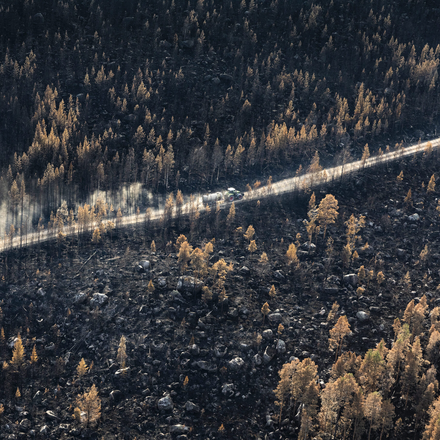 Aerial photo capturing the aftermath of a forest fire in Hälsingland, showing the devastating destruction of the landscape. In the midst of the burnt trees and charred ground, a tractor equipped with a water tank can be seen bravely navigating through the scorched forest, working diligently to extinguish the remaining flames and aid in the recovery efforts.