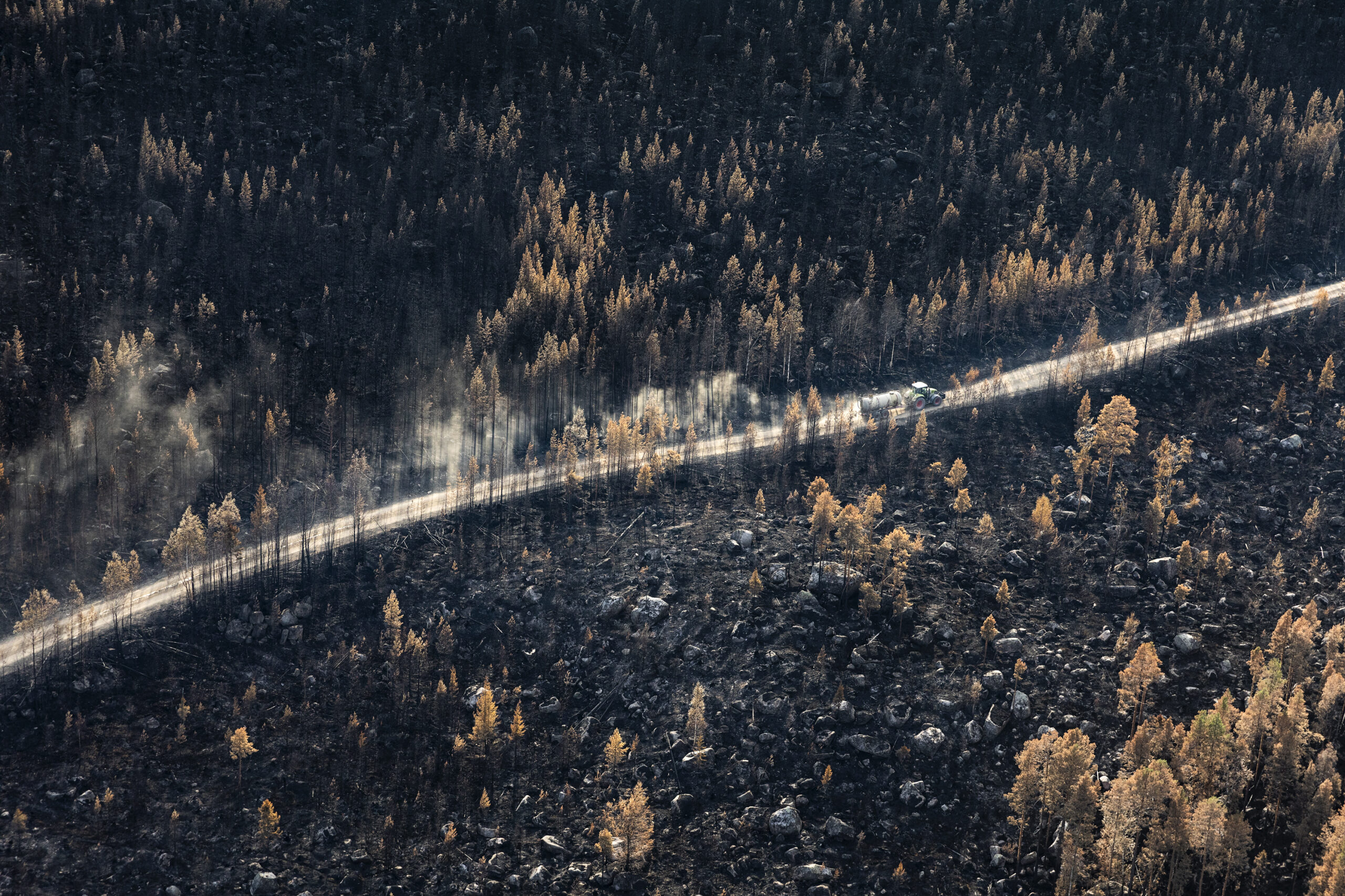 Brand Hälsingland 2_lowres Aerial photo capturing the aftermath of a forest fire in Hälsingland, showing the devastating destruction of the landscape. In the midst of the burnt trees and charred ground, a tractor equipped with a water tank can be seen bravely navigating through the scorched forest, working diligently to extinguish the remaining flames and aid in the recovery efforts.