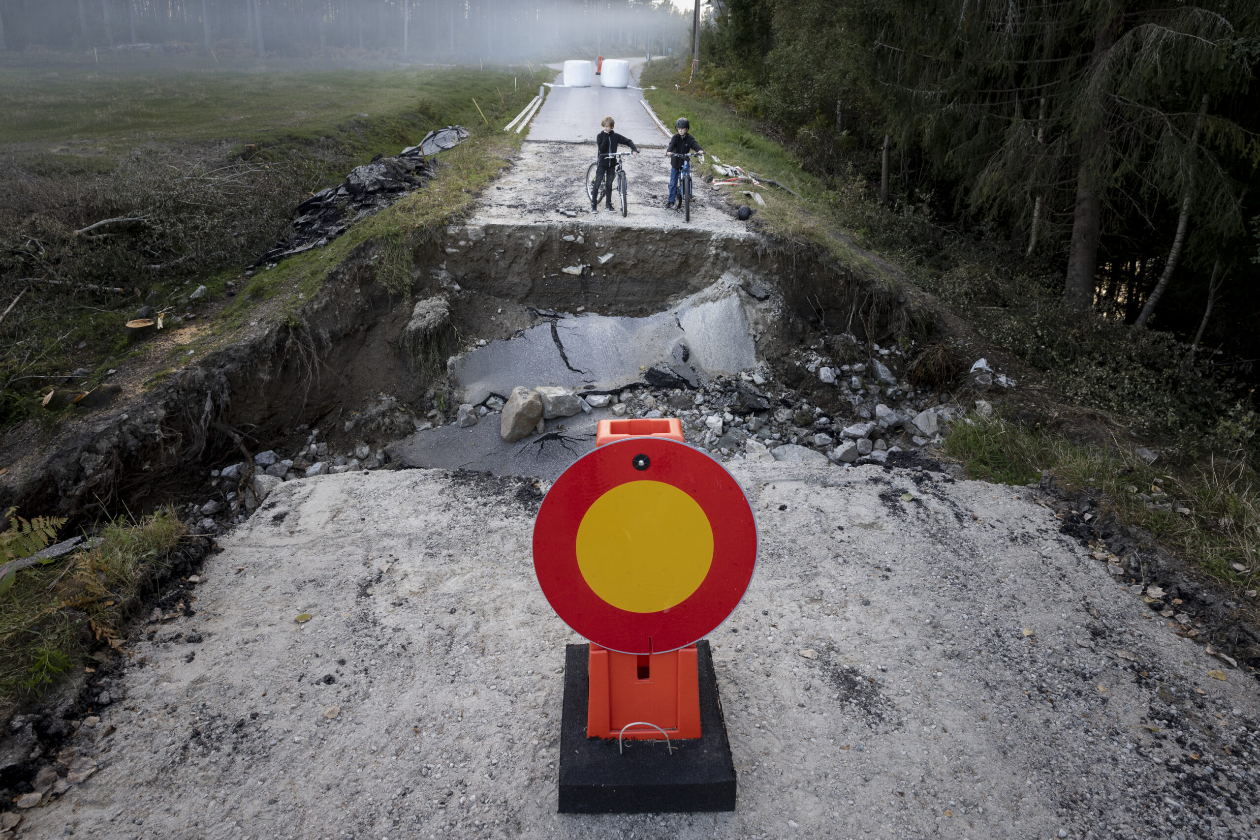 Skyfall Gävle 4 image depicting a collapsed road after the heavy rainfall in Gävle. two boys with their bicycles stand on one side of the collapsed water-damaged road